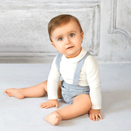 Bébé garçon en chemise blanche et bretelles bleues, assis devant un mur texturé élégant, collection vêtements bébé luxe accessible Chou de Chic.