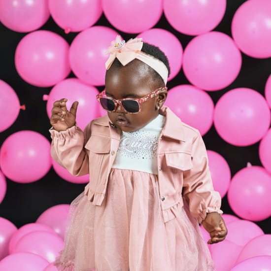 Bébé portant une robe rose avec jupe en tulle, veste assortie, bottines noires et lunettes de soleil, devant un mur de ballons roses, collection vêtements bébé mode d'occasion stylée Chou de Chic.