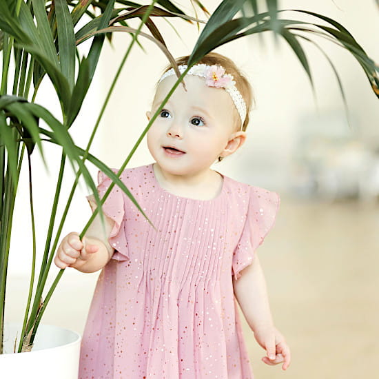 Bébé en robe rose pâle ornée de détails délicats et serre-tête floral, touchant les feuilles d’une plante, collection vêtements bébé chic de seconde main Chou de Chic.