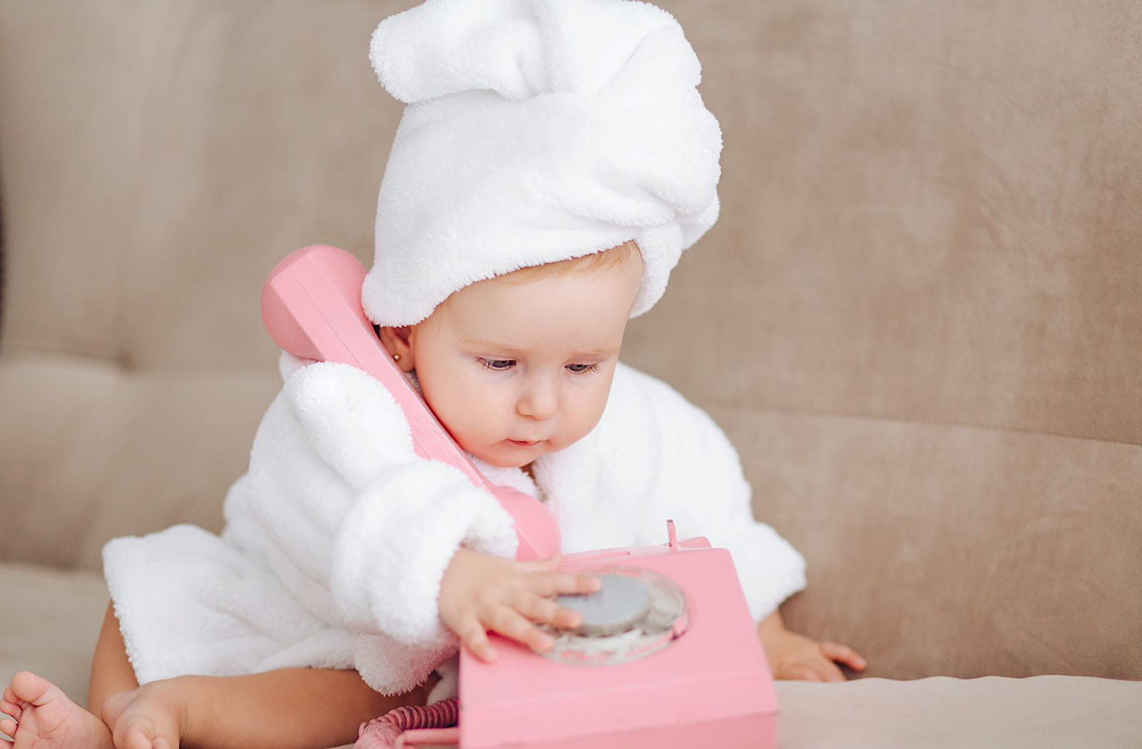 Bébé élégant en peignoir blanc, assis sur un canapé beige, tenant un téléphone vintage rose, illustrant l’élégance seconde main pour bouts de chou